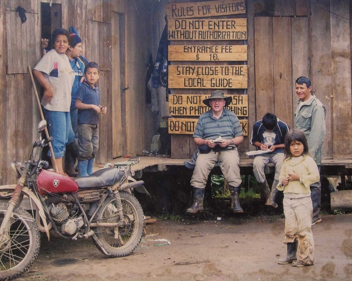 An older man sits on a bench in front of a sign that reads: Rules for visitors; Do not enter without authorization; Entrance fee $10; Stay close to the local guide; Do not use flash when photographing. Family members sit around this gentleman and a motorcycle sits to the right hand side.